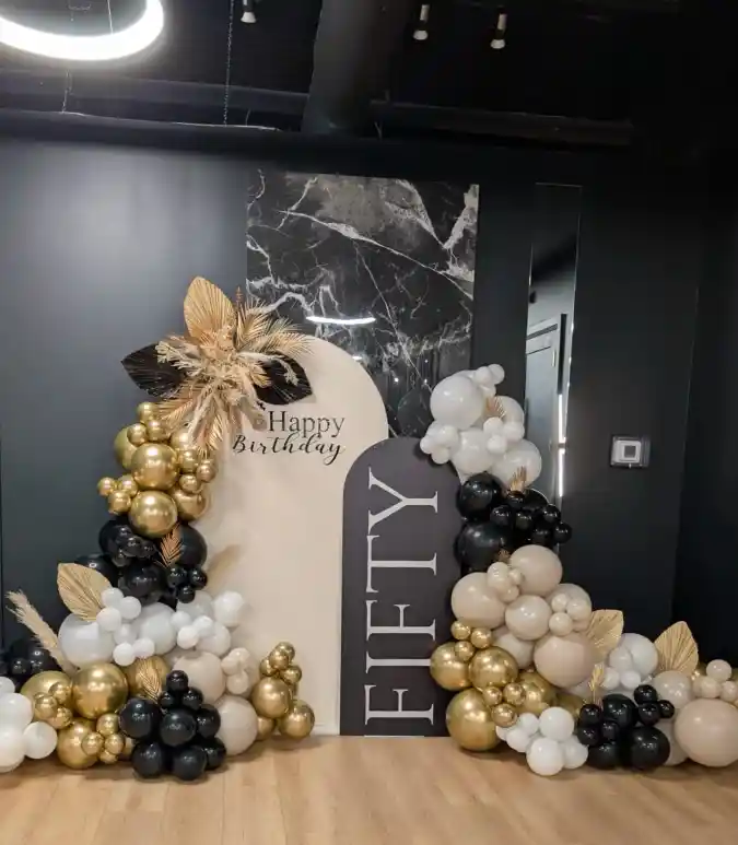 Sophistication defines this stunning fiftieth birthday celebration backdrop, designed specifically to capture memories. A cascading balloon garland featuring metallic gold, matte black, crisp white, and soft nude tones frames the setup elegantly. Textural elements like dried palm spears and fluffy pampas grass intermingle with the globes, adding a modern, organic flair to the arrangement that feels very current.Centrally positioned are two distinct arch-shaped panels creating depth against a dark wall that features a striking marble insert. The lighter beige arch reads "Happy Birthday" in a delicate script, while a bold black panel prominently displays "FIFTY" in large, vertical block letters. This specific configuration provides an excellent focal point where guests can gather to take memorable pictures throughout the night.Executing refined 50th birthday party ideas often involves creating a dedicated, stylish space for photography just like this example. The clever combination of classic color palettes and trendy textures offers a mature yet festive atmosphere suitable for marking half a century. It successfully transforms a simple room corner into a major statement piece honoring the birthday person.